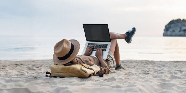An image featuring a person wearing a hat while at the beach and using a laptop.