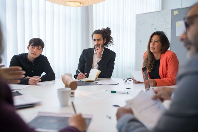 An image showing three people sitting around a table at a working environment and listening to someone carefully.